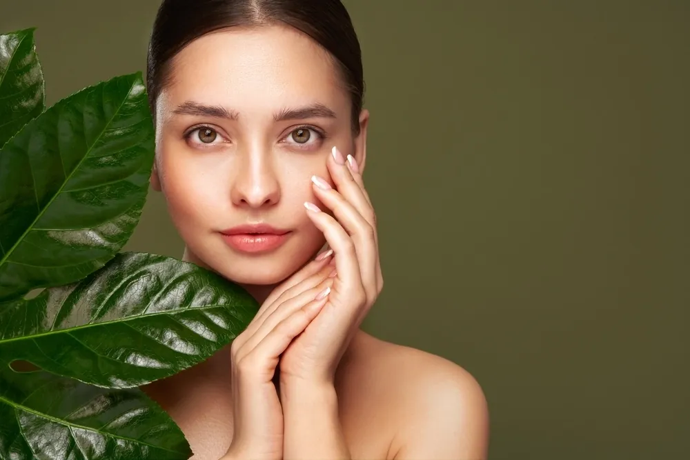 A young woman with clear skin touching her face beside large green leaves