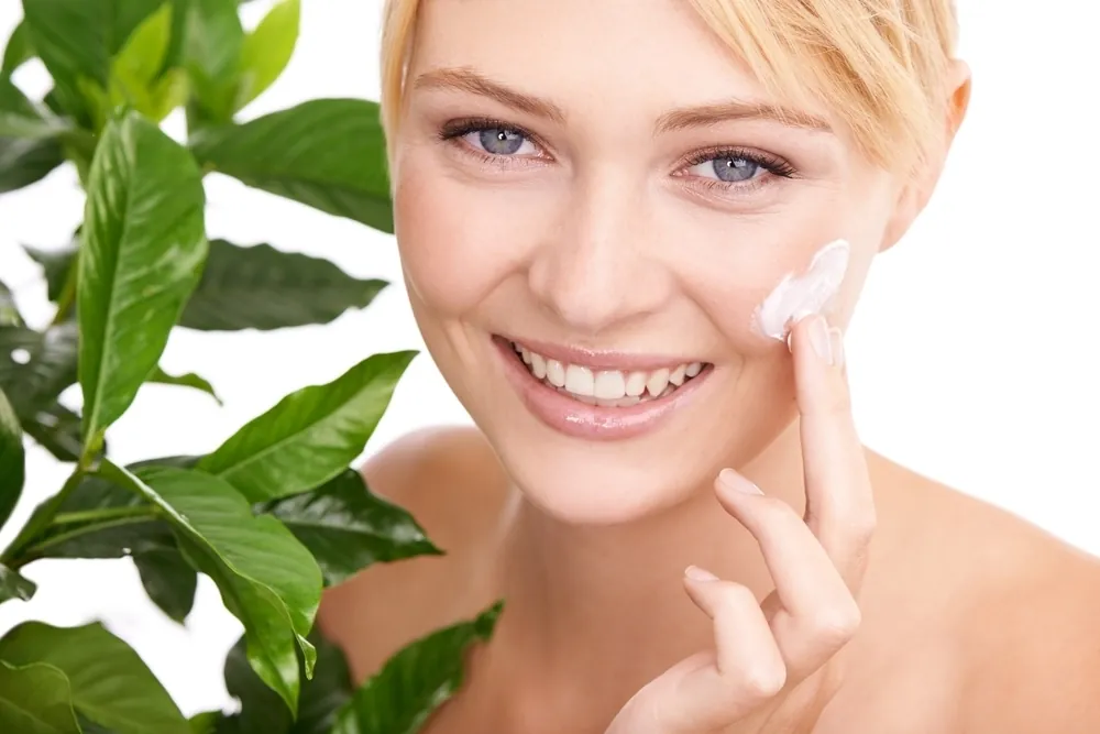 A smiling woman applying cream to her cheek beside green plants