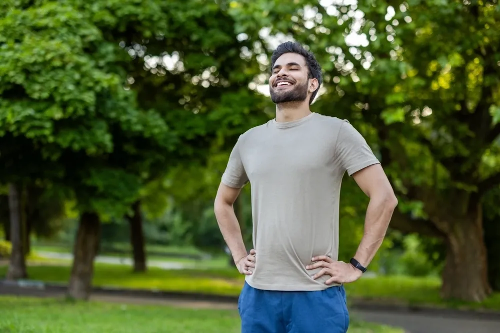 A smiling man standing outdoors in a park with his hands on his hips