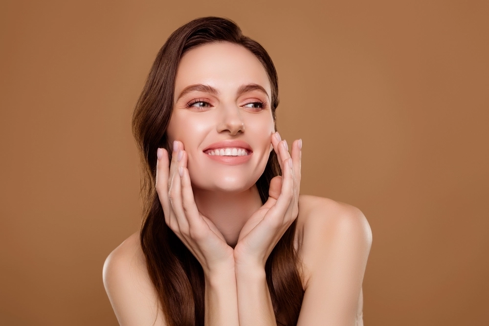 A smiling woman with long brown hair gently touching her face