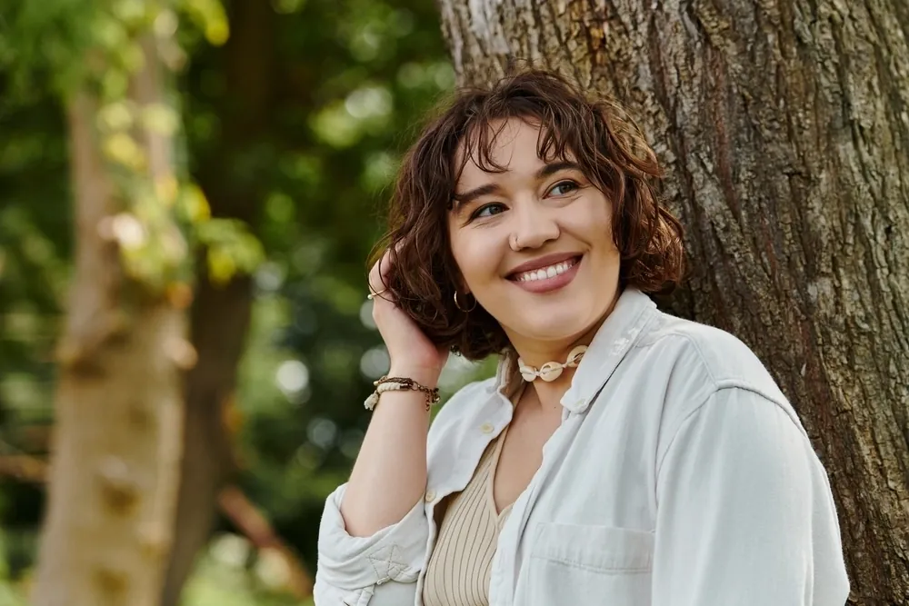 A smiling woman with short curly hair leaning against a tree