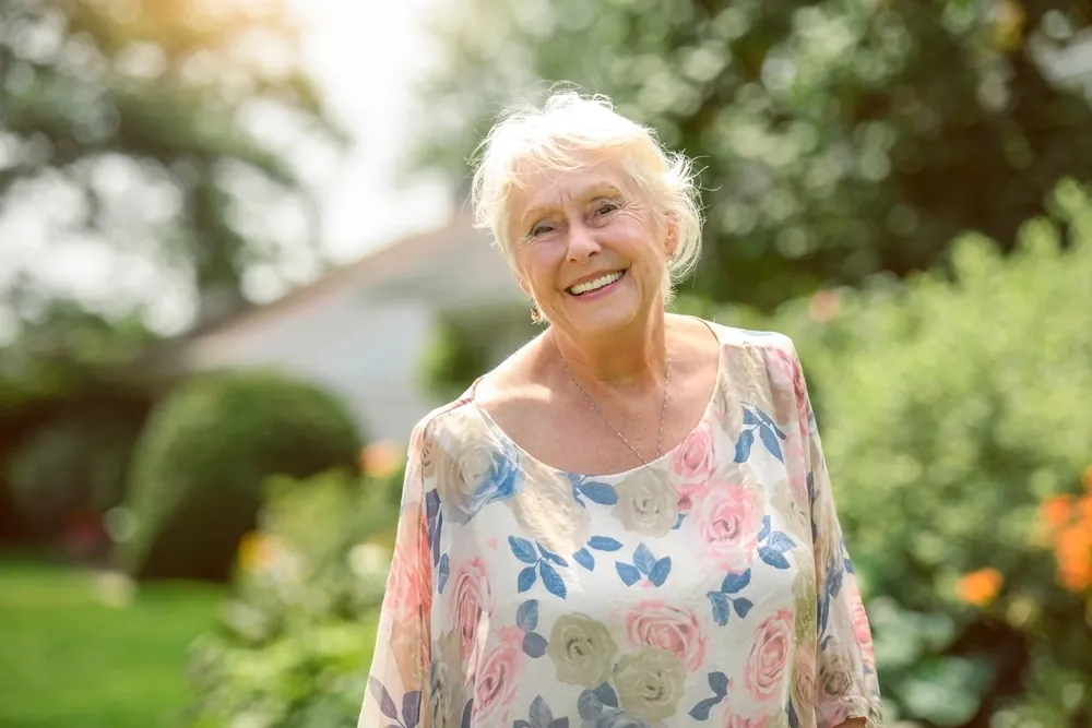 A smiling older woman with short white hair in a sunny garden