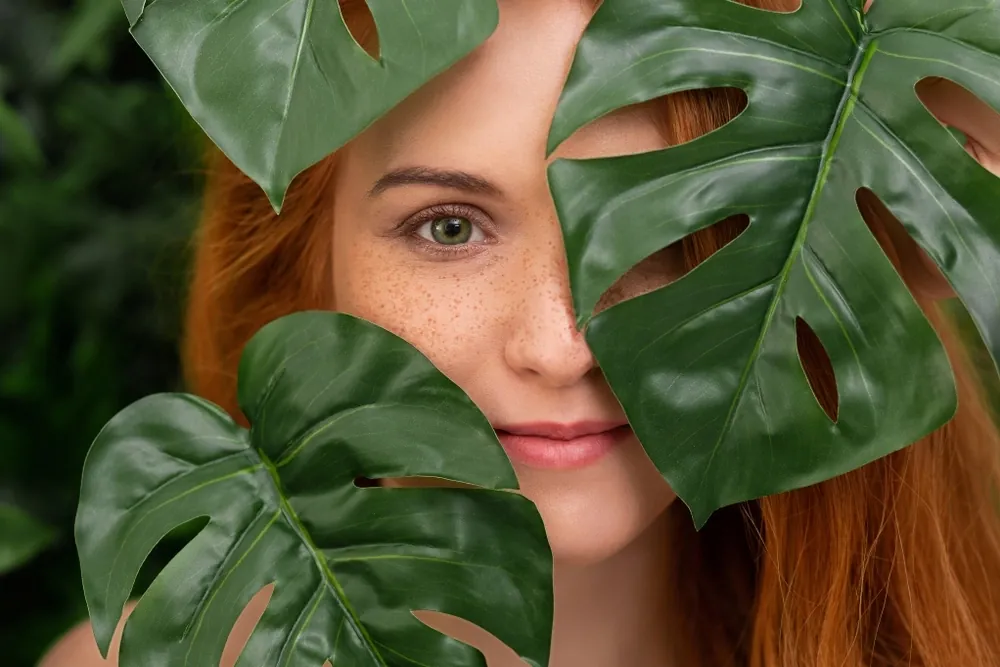 A woman with red hair and freckles partly hidden behind green leaves